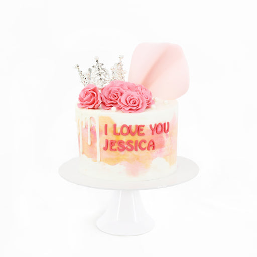 White buttercream frosted cake with pink and orange accents, topped with roses and a crown