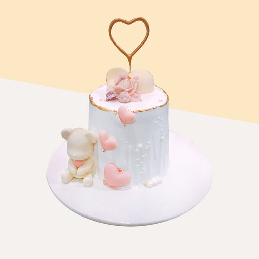White buttercream cake with pink accents, with a teddy bear and heart shaped candle