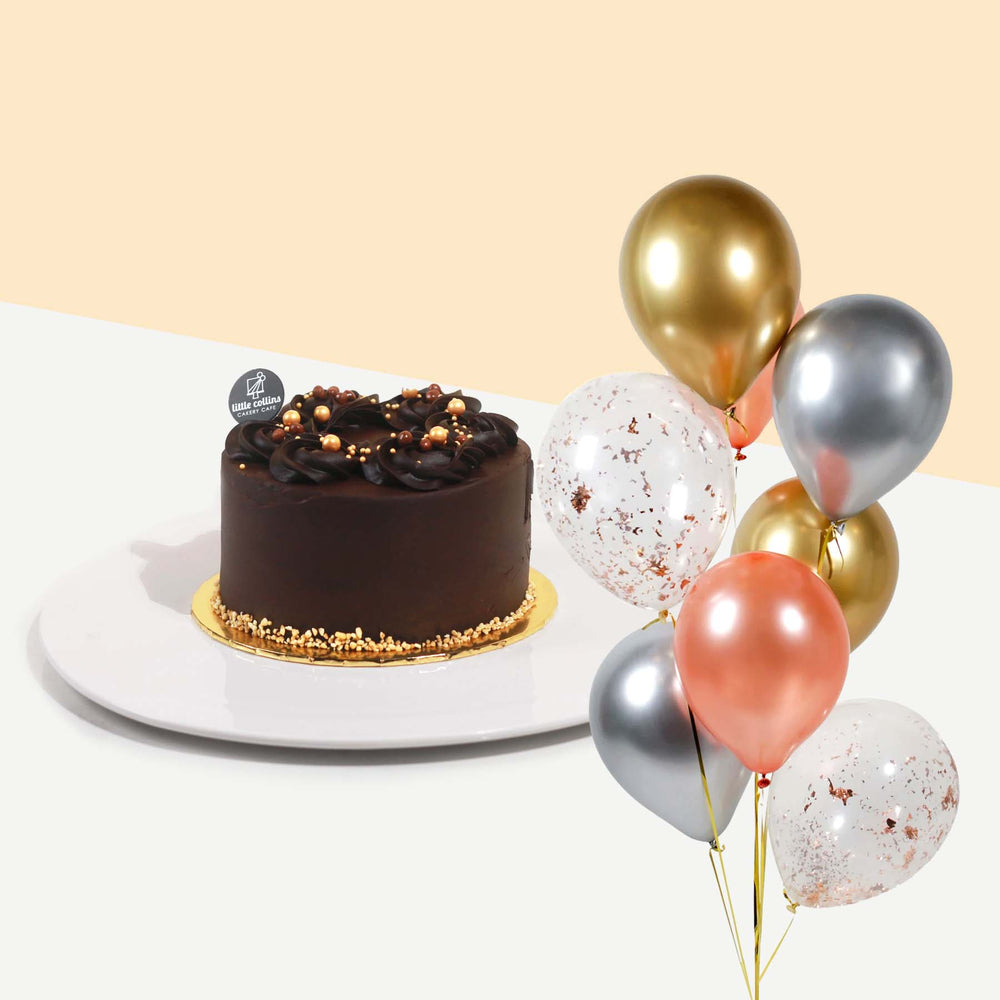 Chocolate cake topped off with edible pearls, beside a bundle of balloons