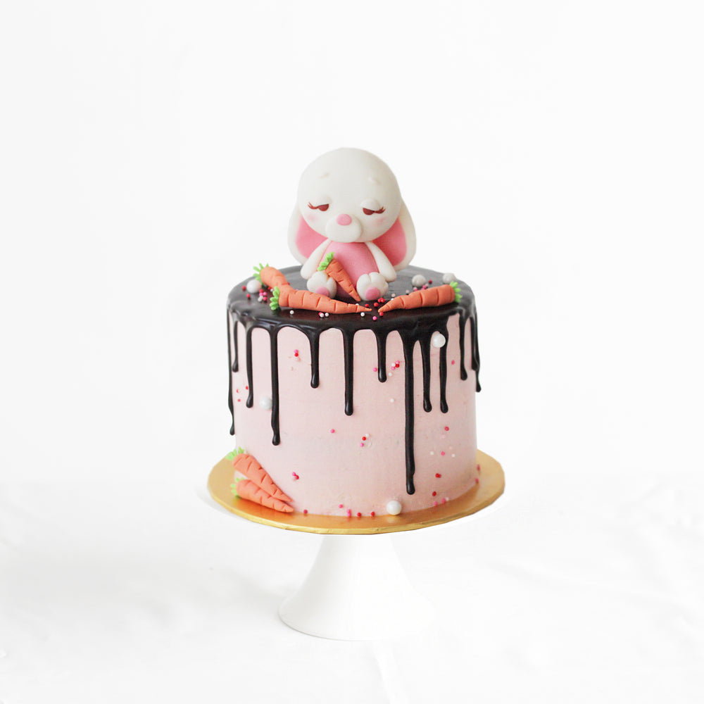 Fondant bunny with fondant carrots, sitting on a butter cake frosted with pink buttercream and chocolate drip