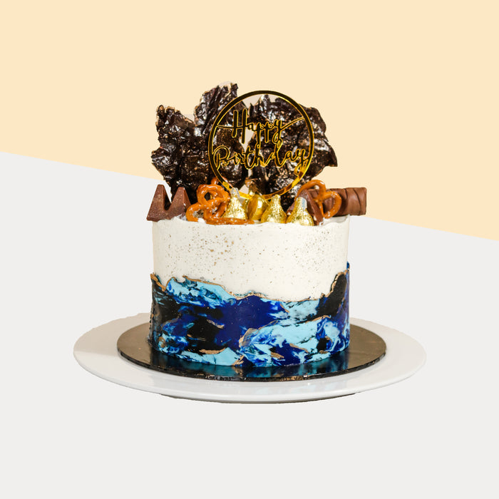 Faultline design cake, with blue marble designs, with while buttercream, topped with pretzels, Kisses and Kinder Bueno