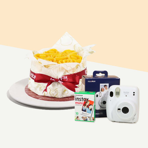 Cake topped with rose shaped mango slices, wrapped in baking paper with ribbon with an Instax camera and film