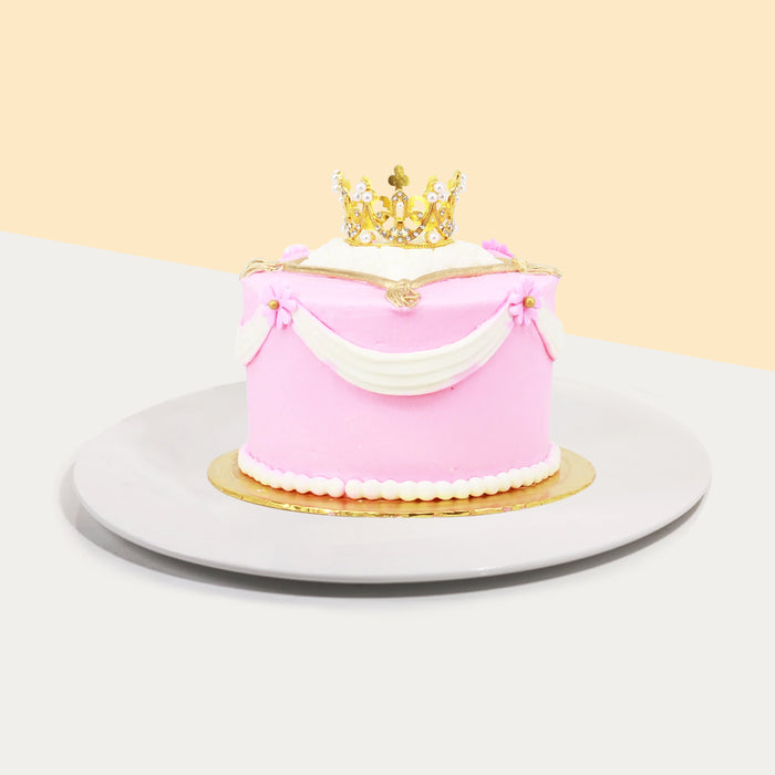Pink princess cake with white buttercream drapes, topped with a golden crown topper